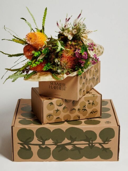 Three stacked cardboard boxes with botanical designs, the top box open and filled with assorted flowers and greenery labeled "SUMMER HARVEST.