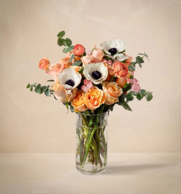A glass vase filled with a bouquet of peach and white flowers, including roses and anemones, with green foliage, set against a beige background.