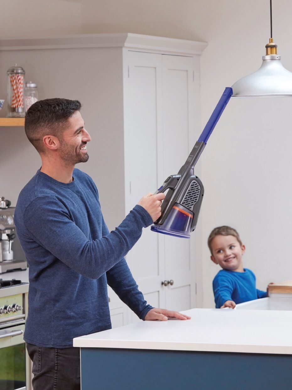 A man uses a cordless vacuum cleaner in the kitchen as a child watches from behind the counter.