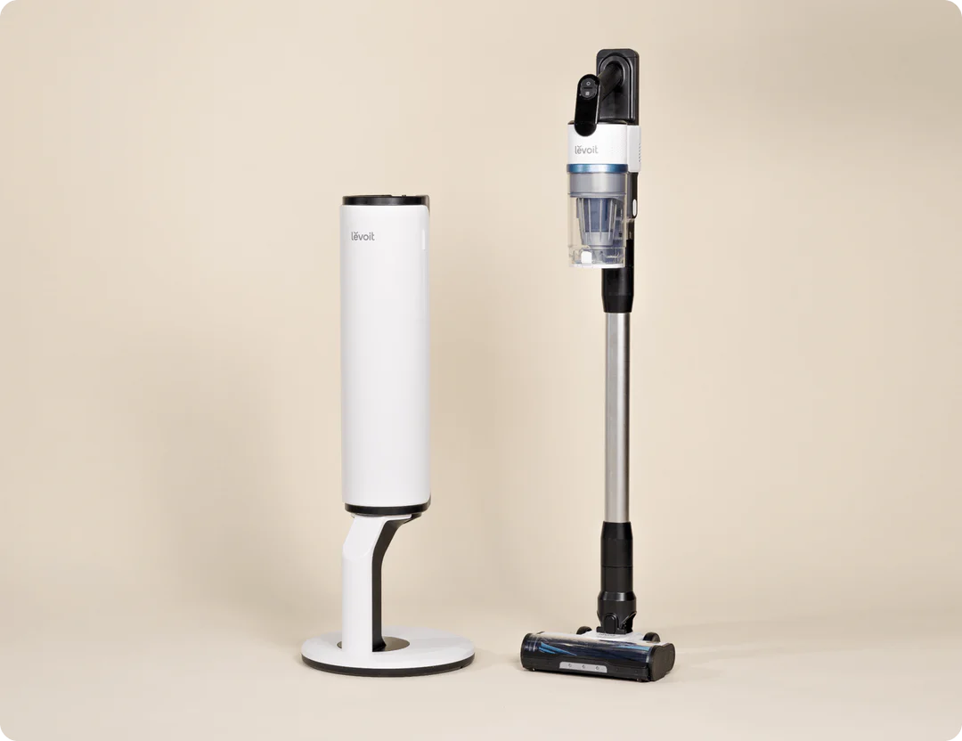 A cordless stick vacuum cleaner stands upright next to its white charging and storage base on a plain beige background.