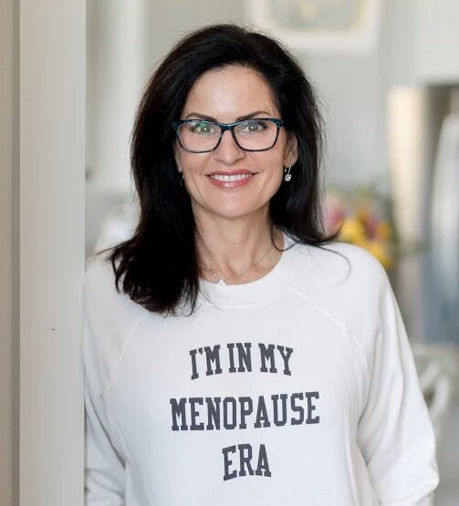 Woman with dark hair and glasses smiling, wearing a white sweatshirt that says "I'M IN MY MENOPAUSE ERA" in bold letters.