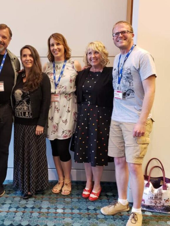 Six adults pose together indoors, wearing conference name tags and lanyards, standing on a blue patterned carpet against a neutral wall.