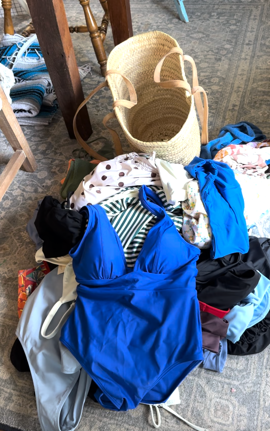 A straw tote bag sits on the floor next to a pile of assorted swimsuits and clothing, with various colors and patterns visible.