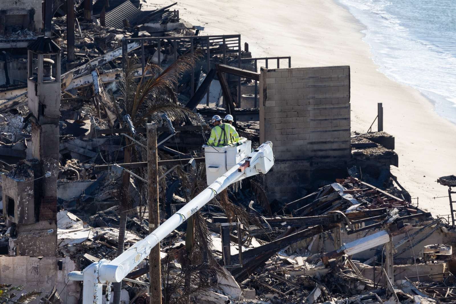 Photo of utility workers in a lift over a background of burned homes and palm trees by the beach