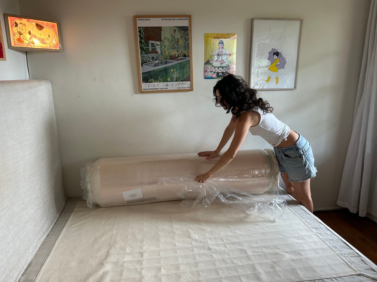A woman in a white tank top and denim shorts unwraps a rolled-up mattress on a bed in a bedroom with framed art on the wall.