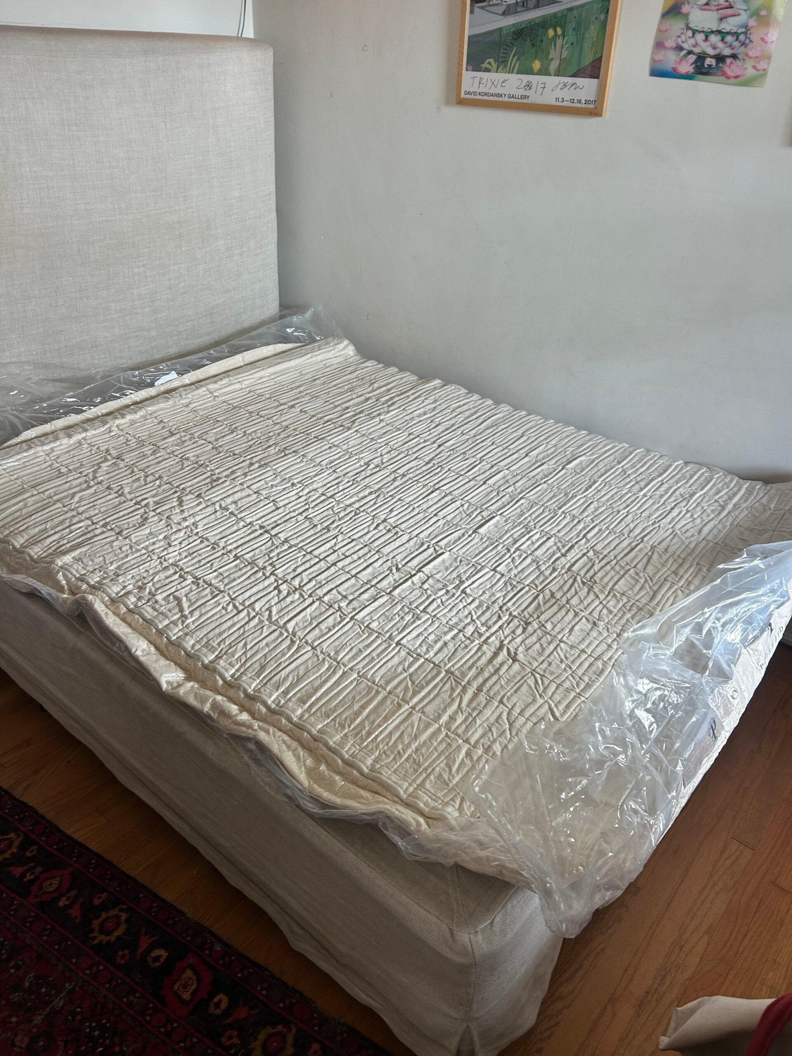 A bed with a light-colored headboard and mattress, partially covered by a protective plastic sheet, stands in a room with framed pictures on the wall.