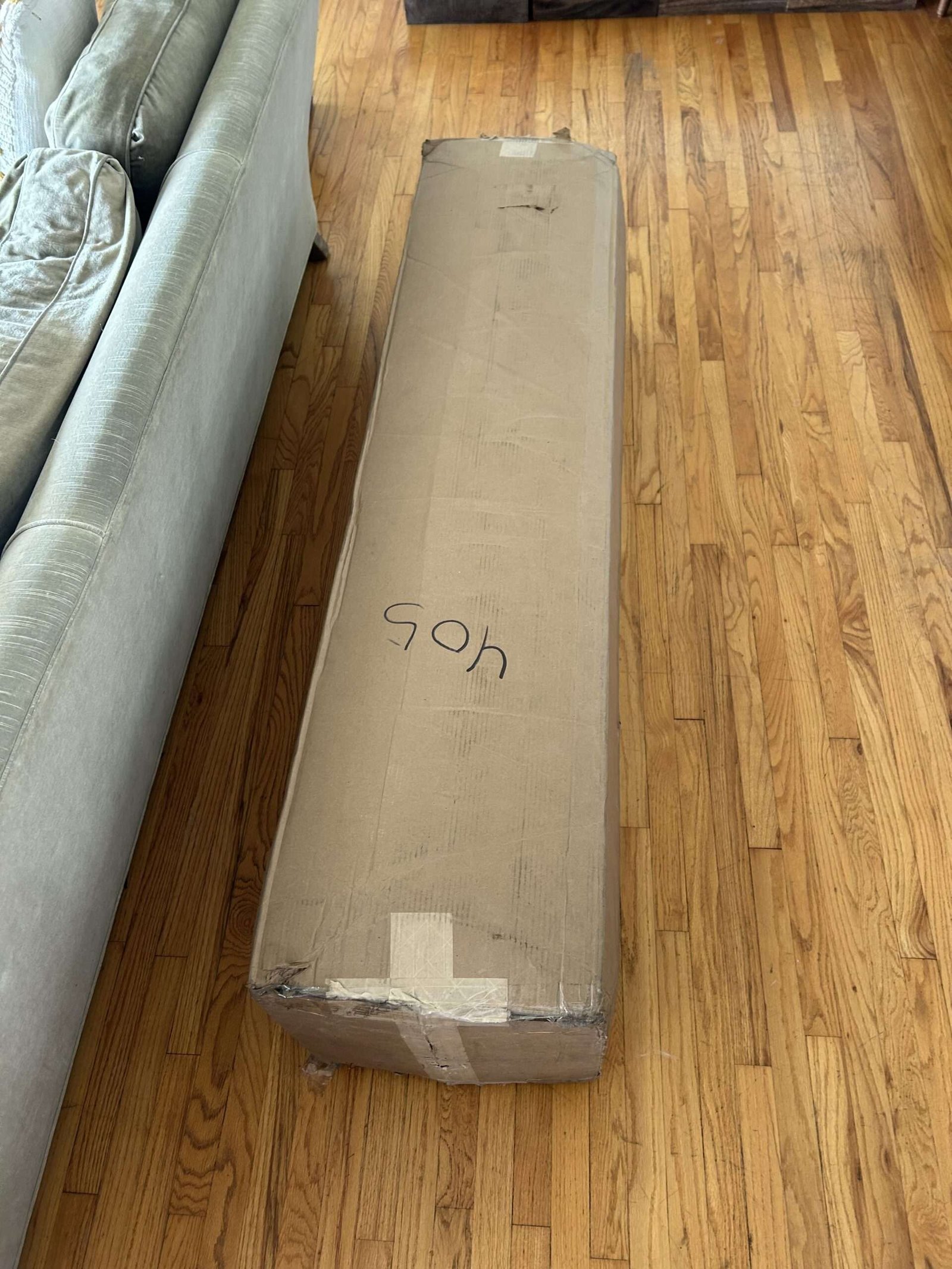 A long, rectangular cardboard box with tape and writing on it sits on a wooden floor next to a green couch.