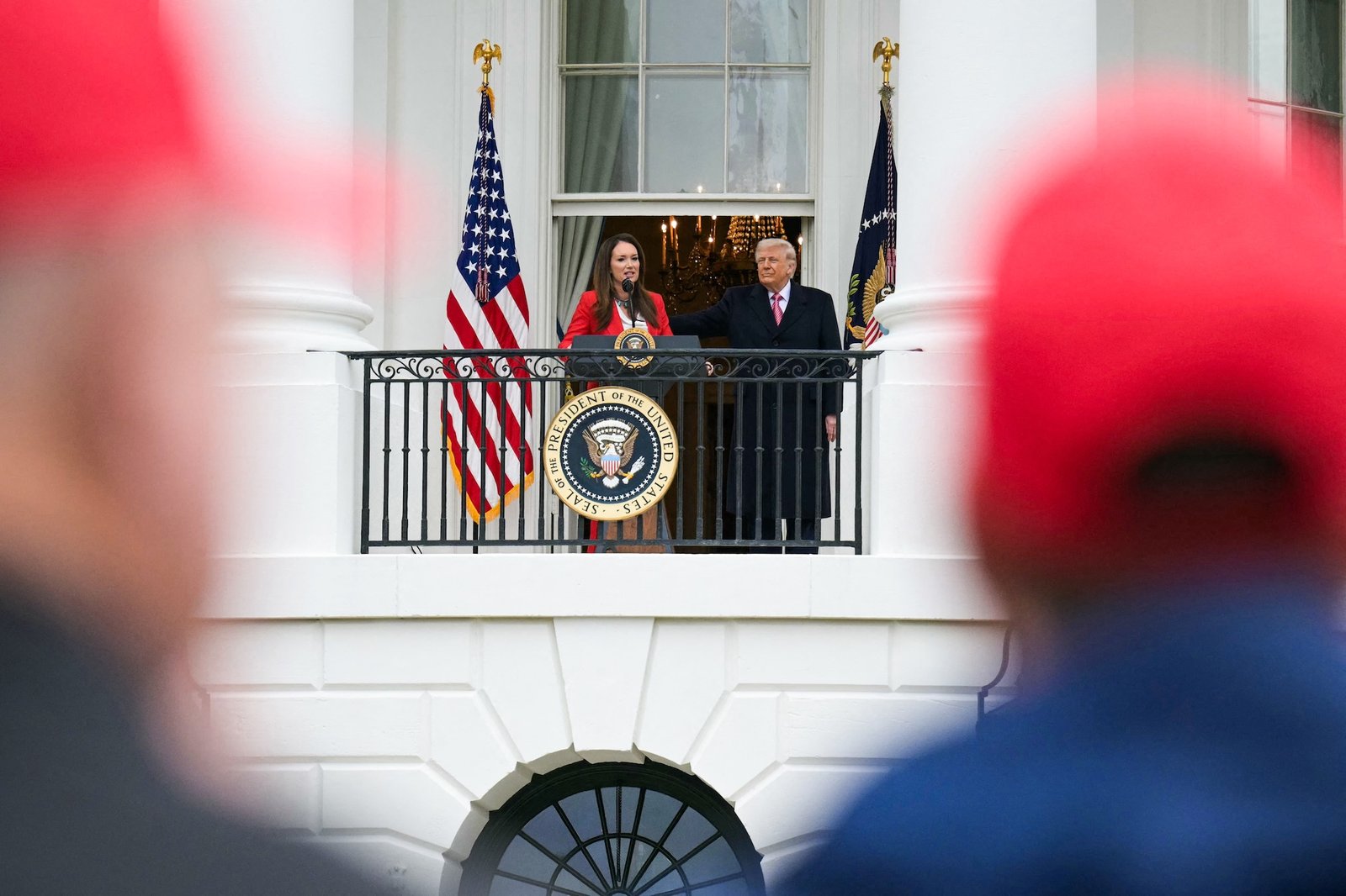 trump and rollins stand on a balcony with the presidential seal on it, as seen from among a crowd of people wearing red hats