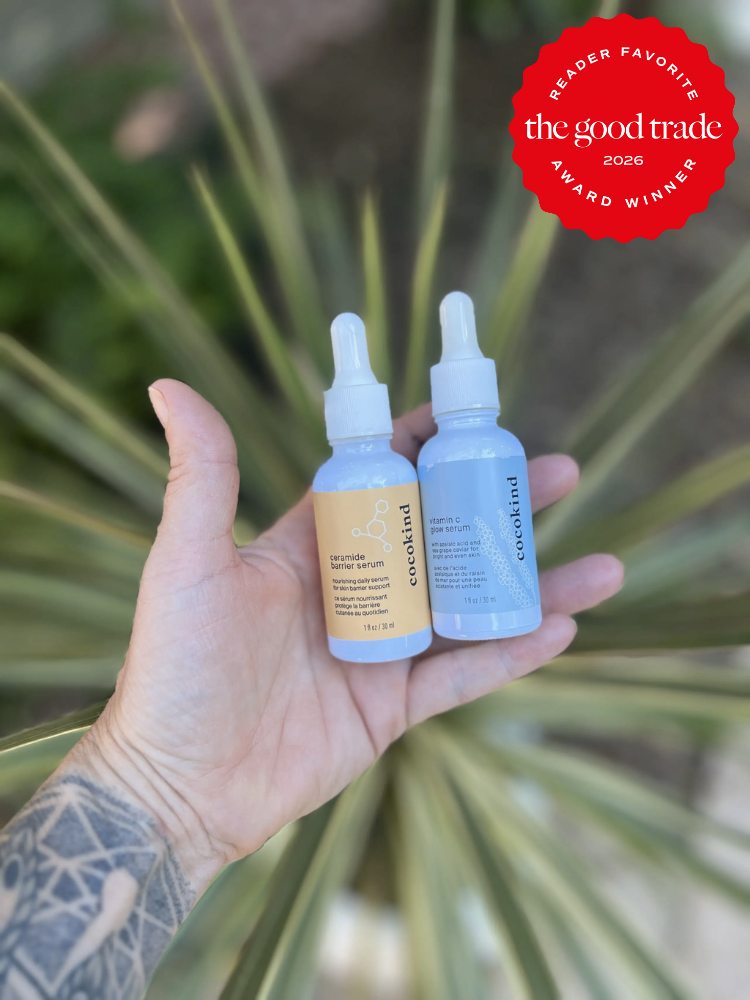 A tattooed hand holds two dropper bottles of Cocokind skincare serum outdoors, with a "The Good Trade 2026 Award Winner" badge in the top right corner.
