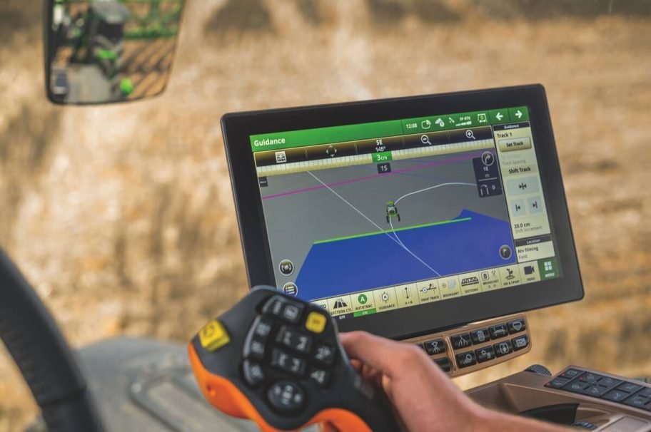 A close-up of a John Deere in-cab display showing a green field-guidance map, with an operator's hand resting on the control console. Photo: John Deere