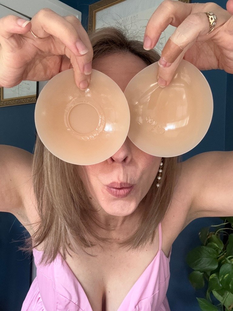Woman holds two nude-colored silicone bra inserts over her eyes and puckers her lips, posing playfully in a pink top.