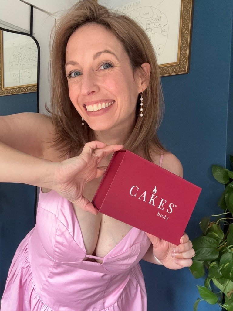 Woman in a pink dress smiling and holding a red box labeled "CAKES body" in front of her, standing indoors next to a plant.