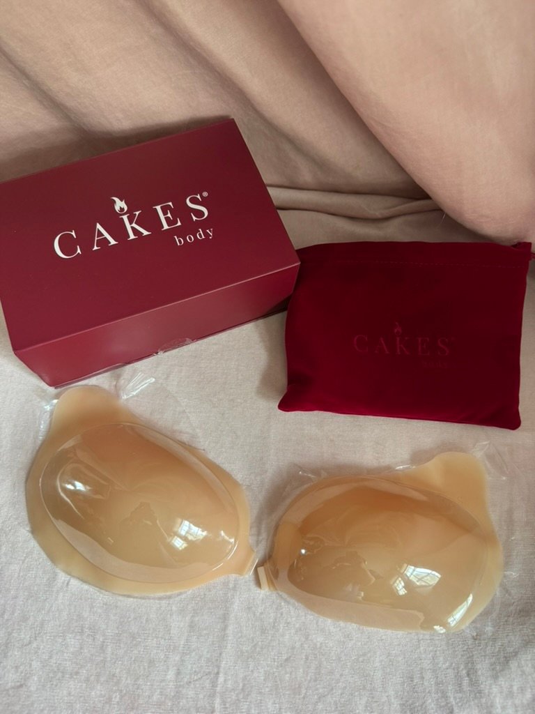 Two adhesive silicone bra inserts are displayed on a surface next to a red CAKES body box and a red storage pouch.