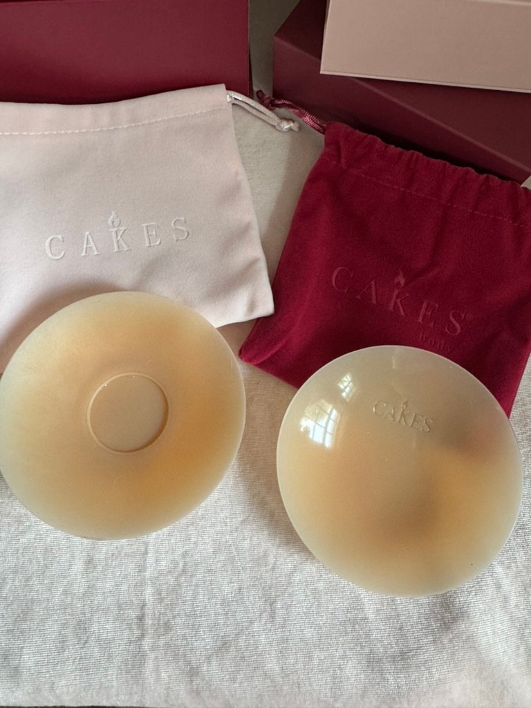 Two round, beige silicone nipple covers are displayed on a white surface, with pink and red CAKES branded drawstring storage bags in the background.