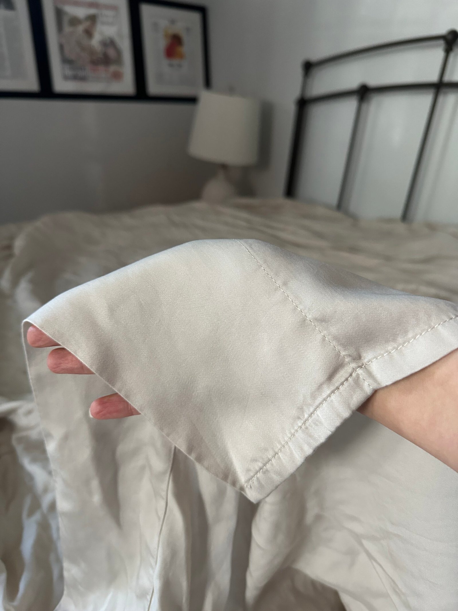 A hand holding the corner of a light-colored bedsheet with visible stitching, in a bedroom with a lamp and framed pictures on the wall.