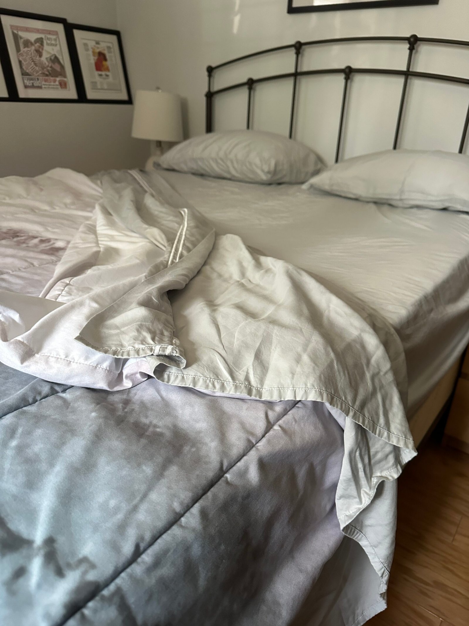 Unmade bed with white sheets and pillows, one bedside lamp, and framed pictures on the wall above a metal headboard.