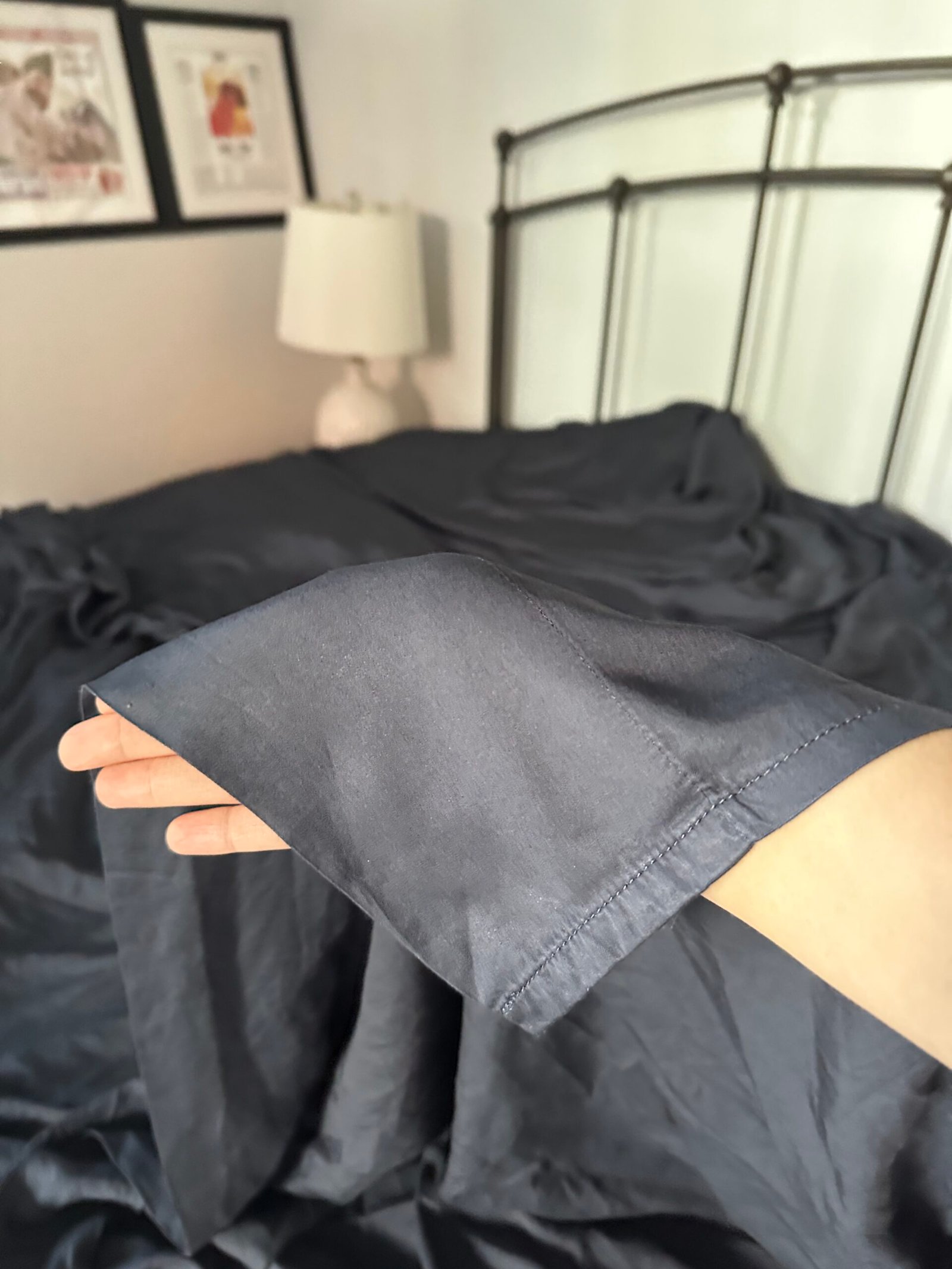 A hand holds the corner of a dark gray bedsheet in front of an unmade bed with a metal headboard, lamp, and framed art on the wall.