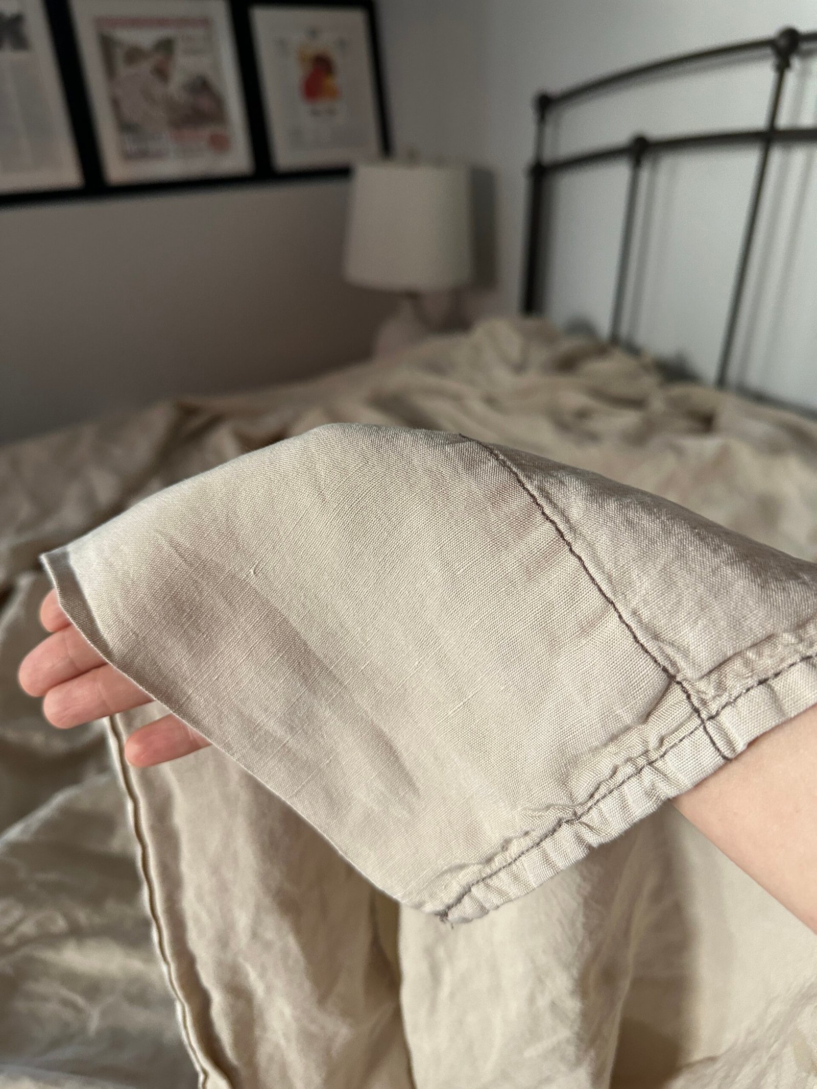 A hand holds up the corner of a beige linen blanket in a bedroom with framed pictures, a lamp, and a metal bedframe visible in the background.