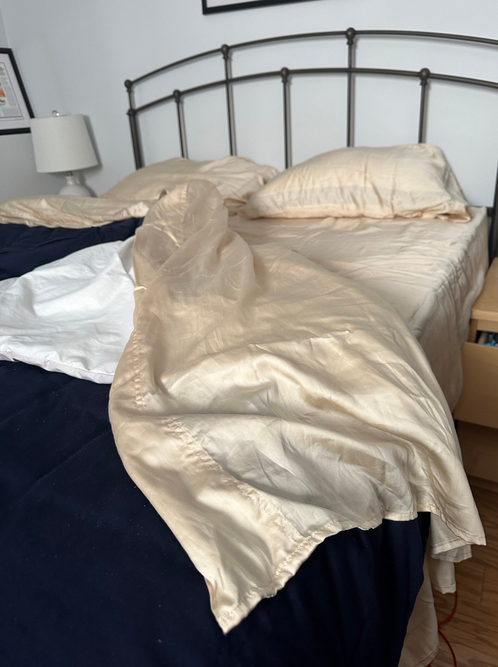 Unmade bed with cream-colored sheets and pillows, a black comforter, and a metal headboard. A lamp and nightstand are visible beside the bed.