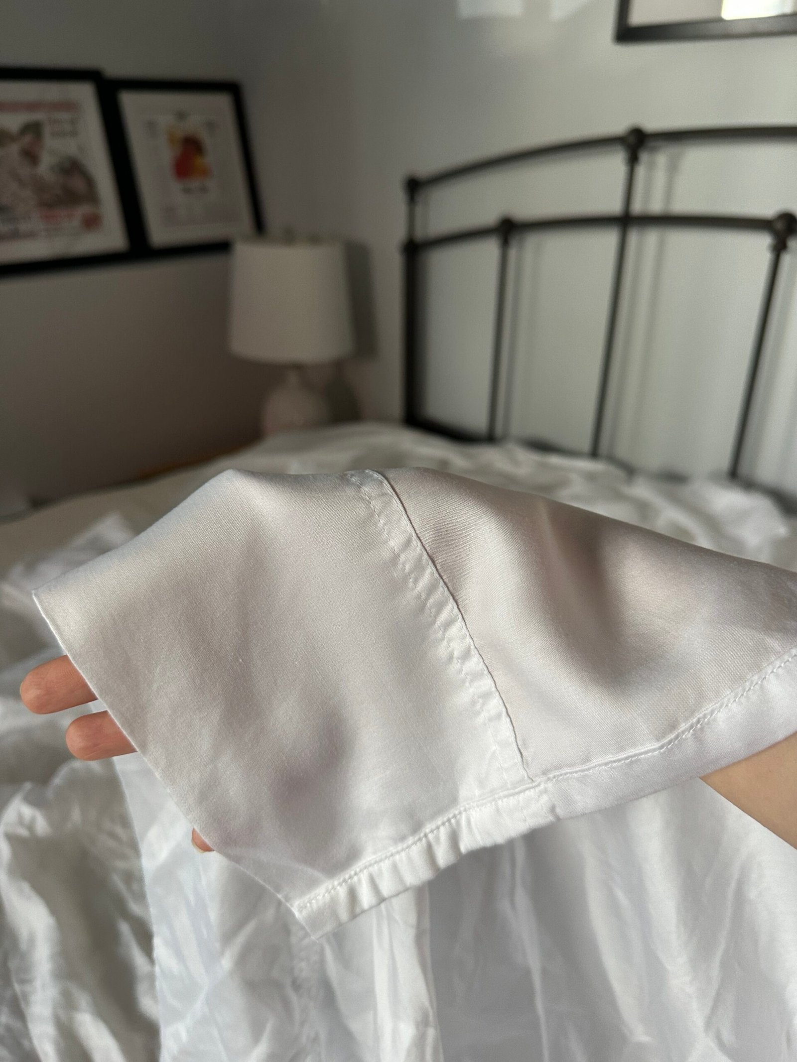 A hand holds up the cuff of a white satin long-sleeve shirt or pajama top against a bed and lamp in a softly lit bedroom.