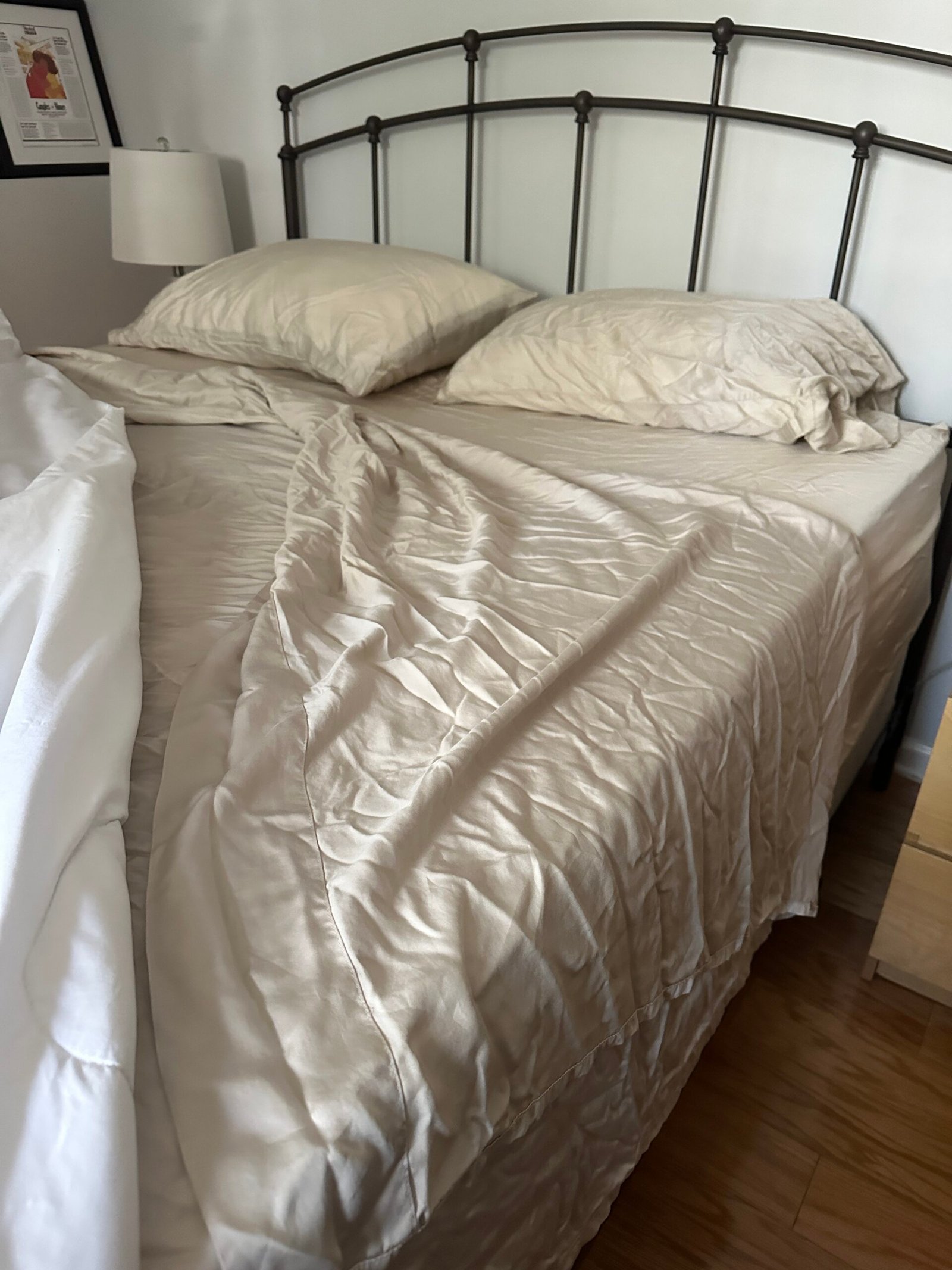 A queen-sized bed with beige sheets and two pillows, partially unmade, sits next to a nightstand and a lamp, on a wooden floor.