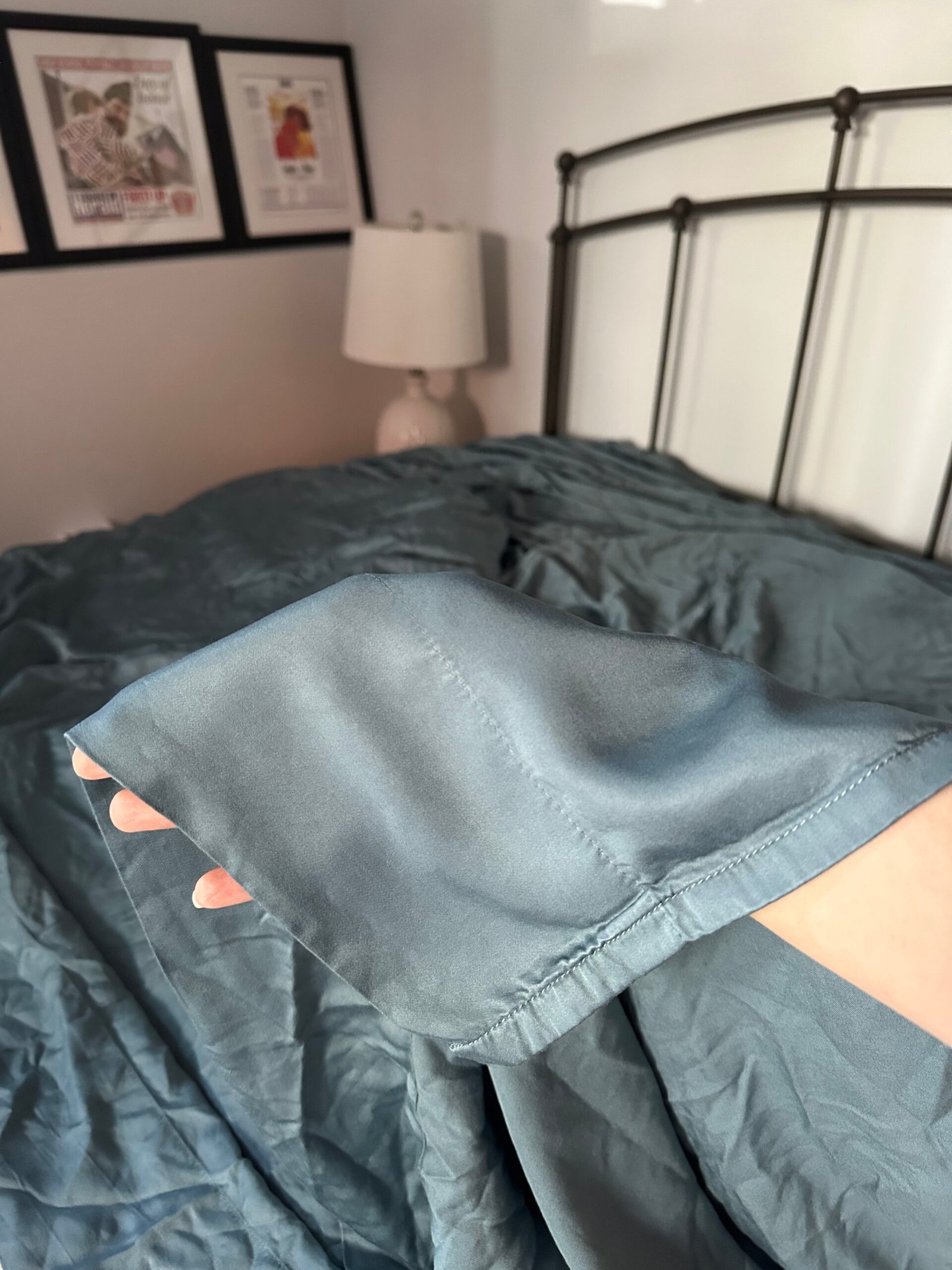 A hand holds up the corner of a wrinkled, teal bed sheet on a made bed with a metal headboard, lamp, and framed pictures in the background.
