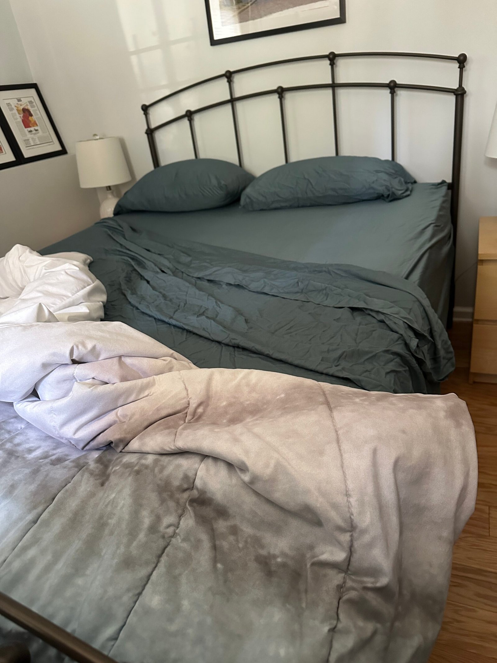 A bed with dark green sheets and two pillows, partially unmade, with a light gray blanket draped messily over the edge and onto the floor.