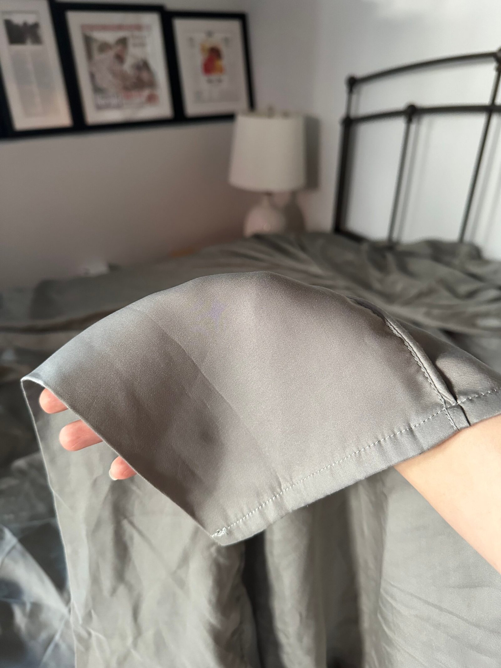 A hand holds the edge of a gray satin sheet in a bedroom with a bed, white lamp, and framed pictures on the wall in the background.