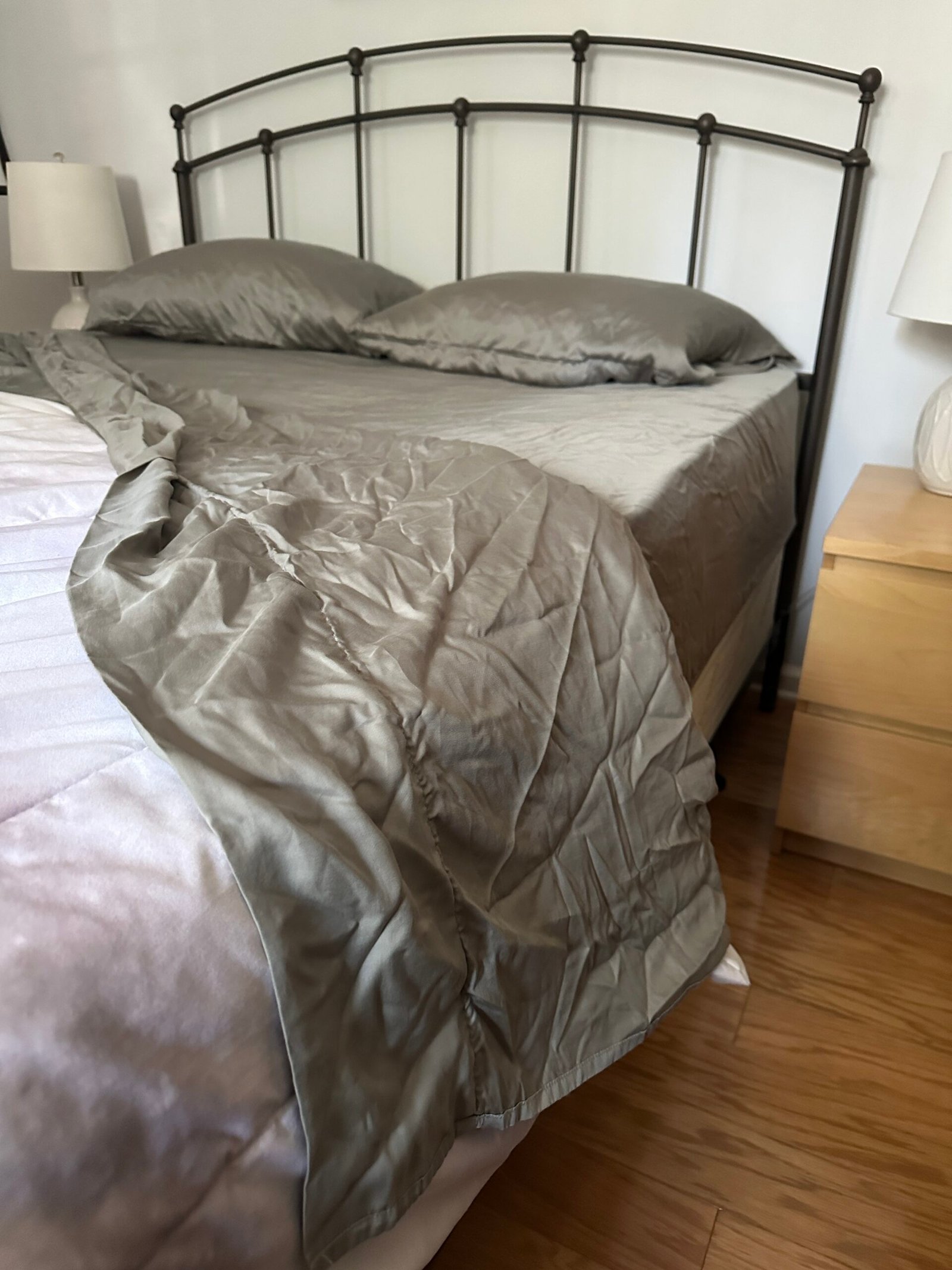 A neatly made bed with gray sheets and pillows, one corner of the blanket is pulled back, revealing a wooden floor and a bedside table with a lamp.