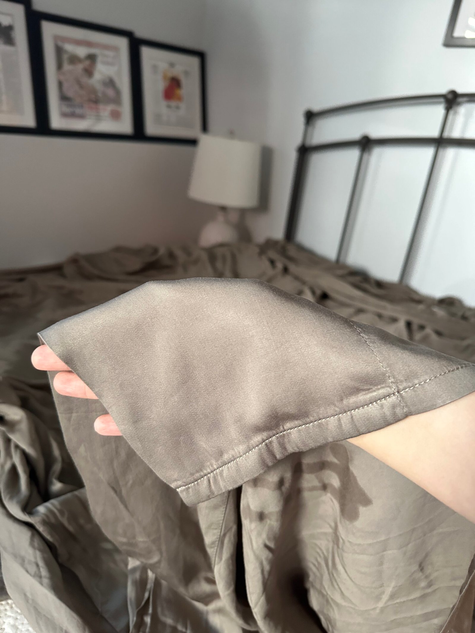 A hand holds up the corner of a taupe satin bedsheet in a sunlit bedroom with framed art on the wall.