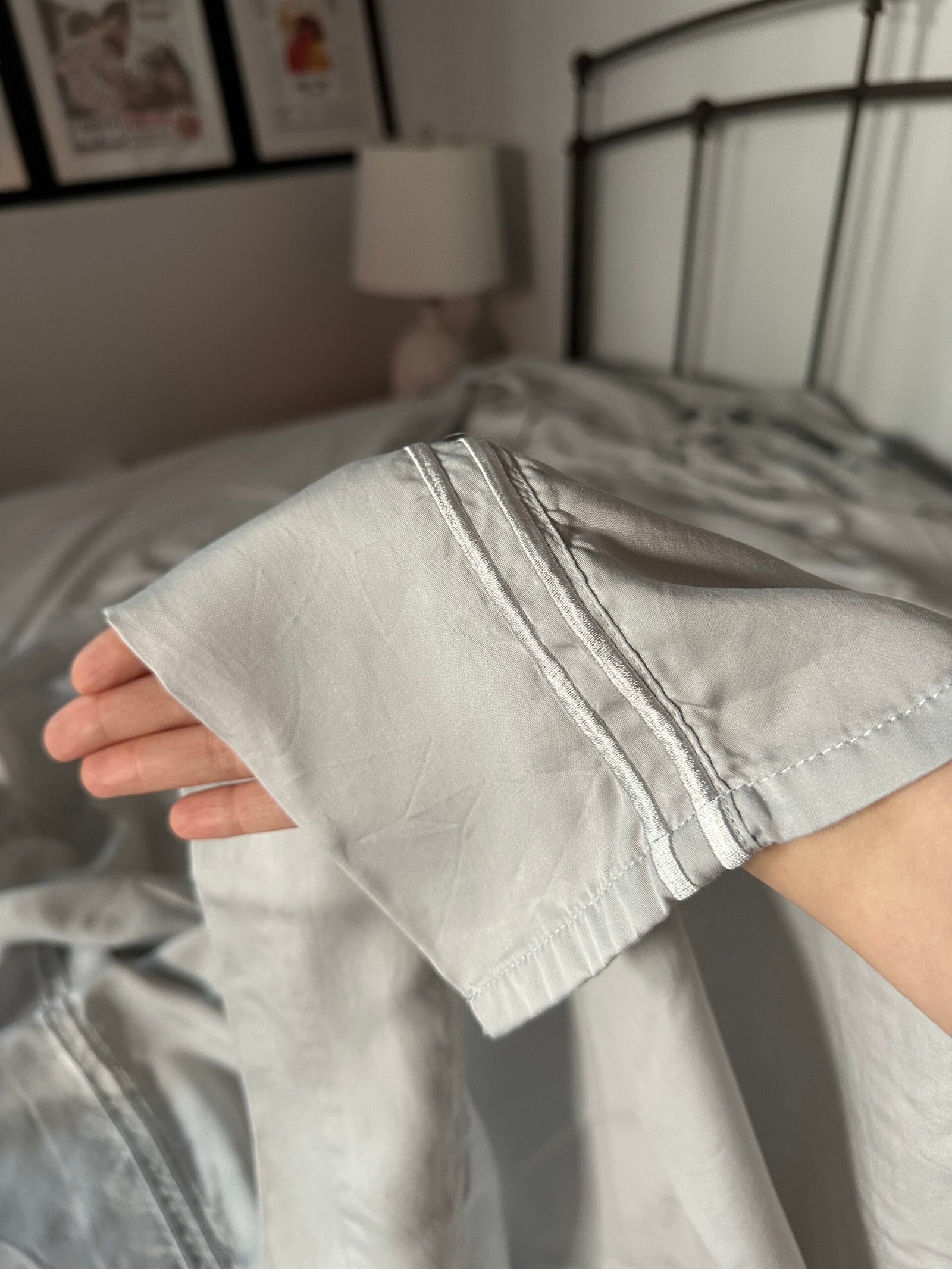 A hand holds up the corner of a light gray bedsheet in a bedroom with a lamp, metal headboard, and framed pictures on the wall.