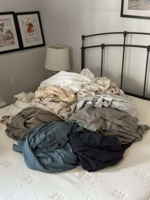 A pile of assorted colored bedsheets and linens is spread across a neatly made bed in a bedroom with framed pictures on the wall.