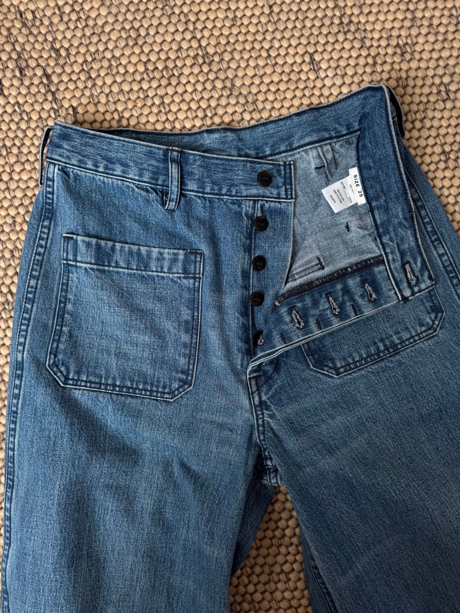 A pair of blue jeans with a button fly partially open, displayed on a textured beige surface.