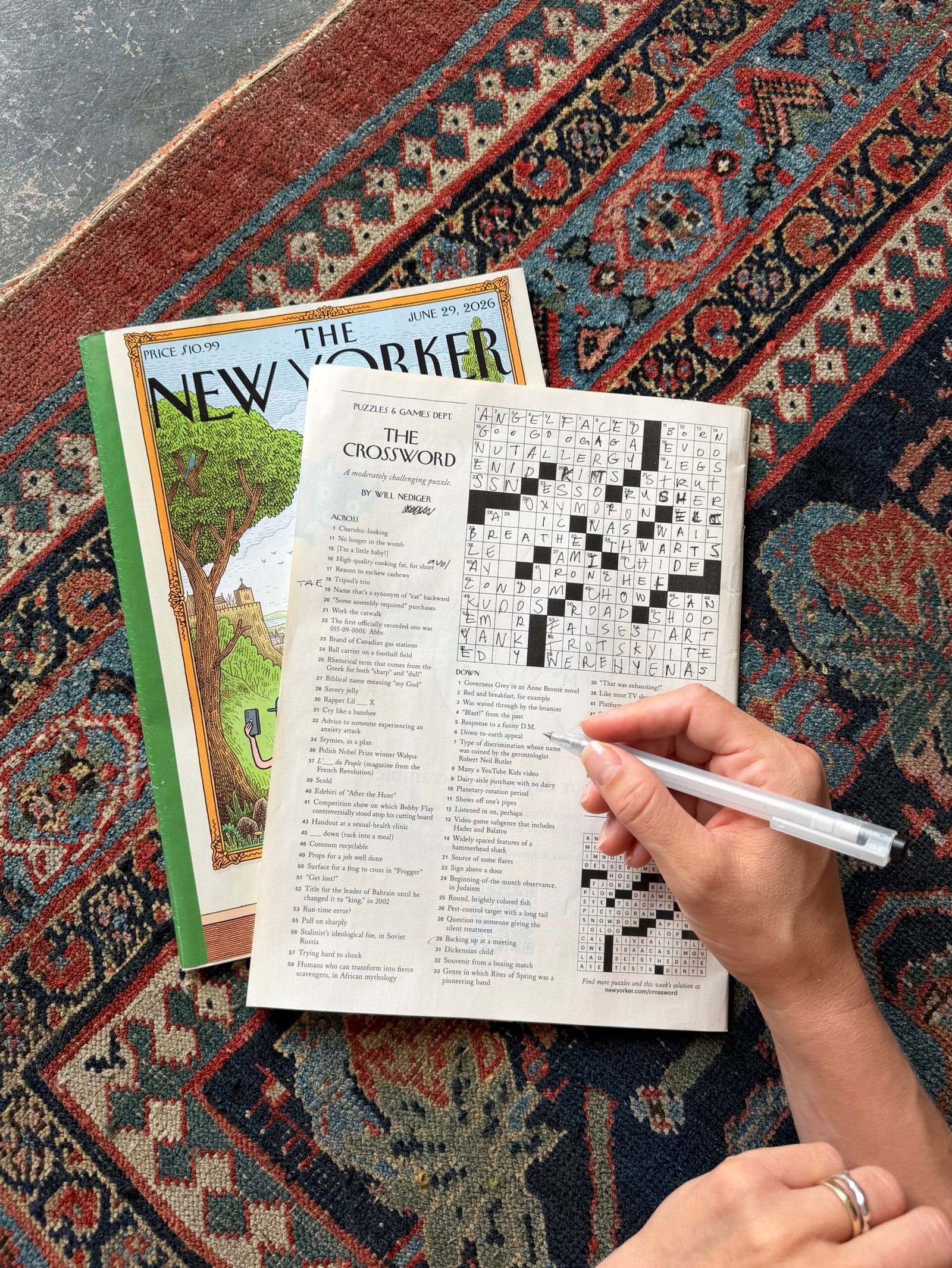A person completes a crossword puzzle in a New Yorker magazine while seated on a patterned rug; another issue of the magazine is partially visible beneath.