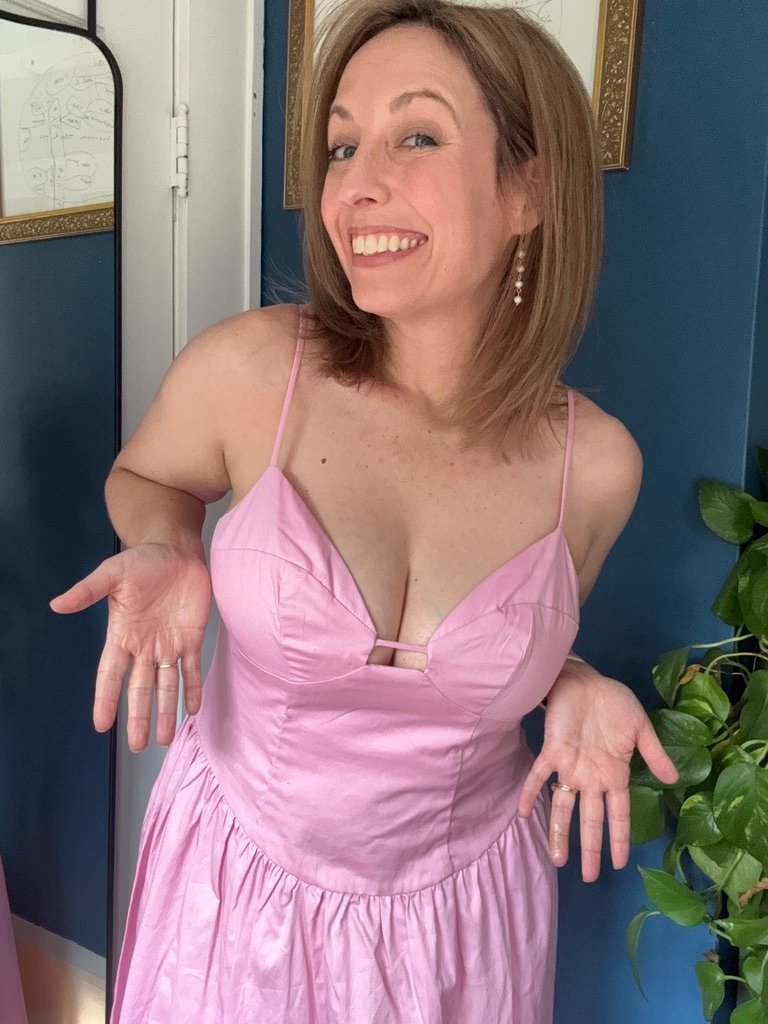 Woman in a pink dress with spaghetti straps stands indoors, smiling and shrugging with palms up.
