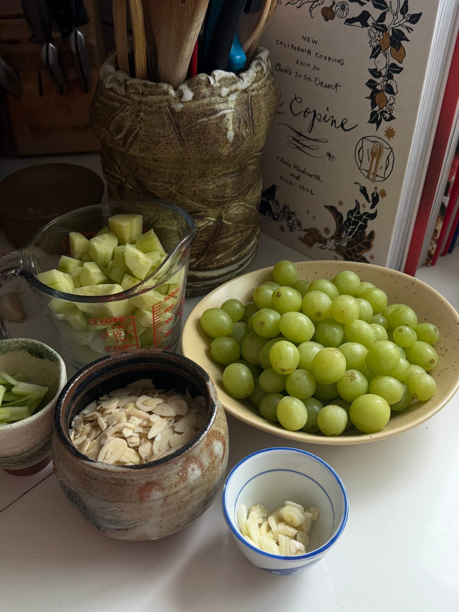 A bowl of green grapes, chopped cucumber, sliced almonds, and garlic are arranged on a counter near a cookbook and utensils.
