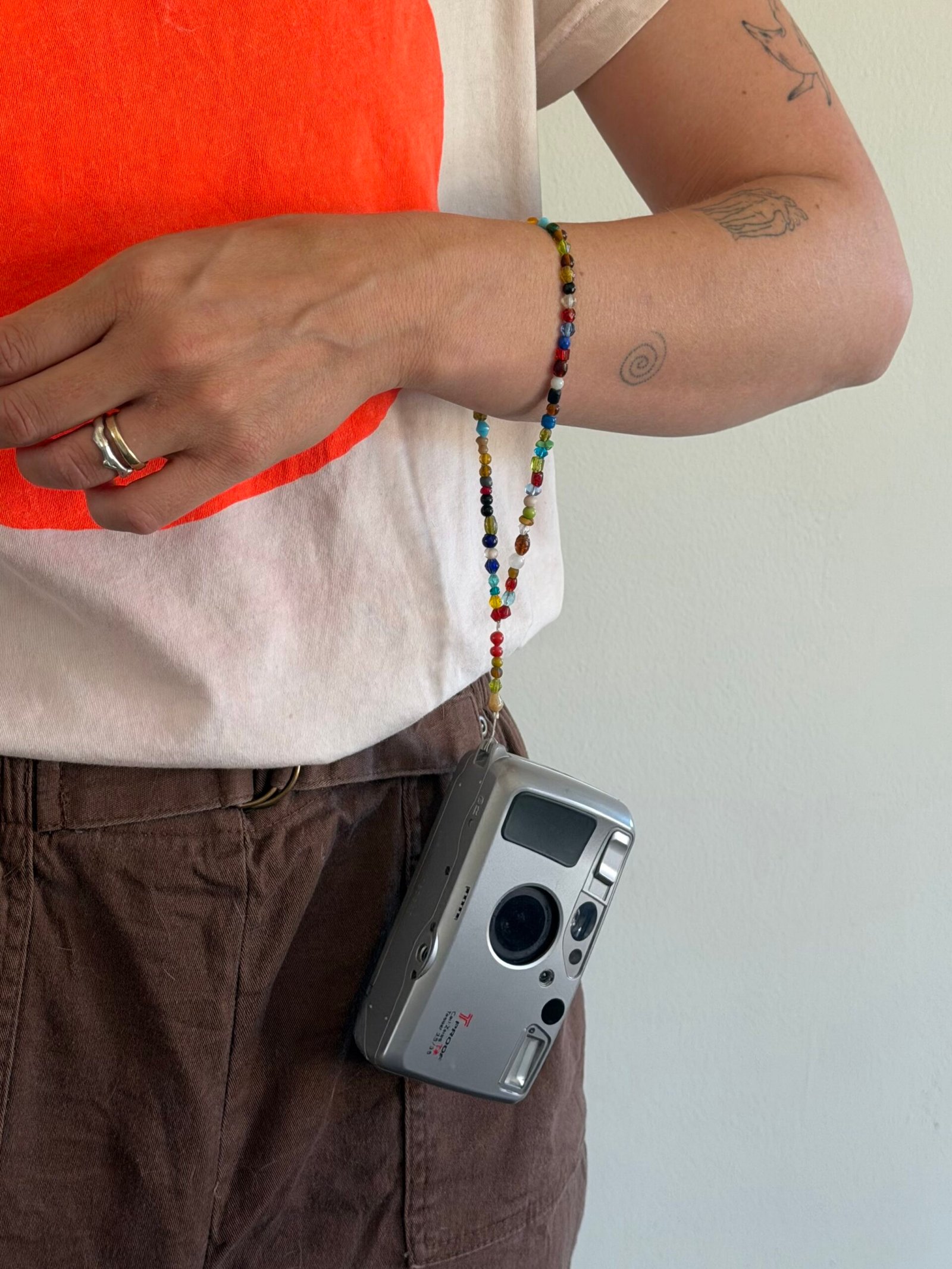 Person wearing a white and red shirt and brown pants holds a silver film camera with a beaded strap. Visible arm tattoos and two rings on their hand.