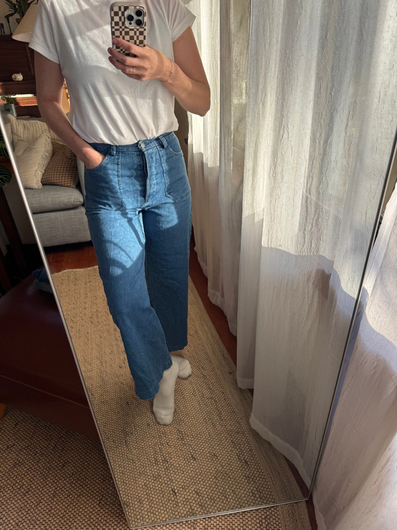 Person taking a mirror selfie, wearing a white t-shirt, blue high-waisted jeans, and white socks, standing on a woven rug in a sunlit room with light curtains.