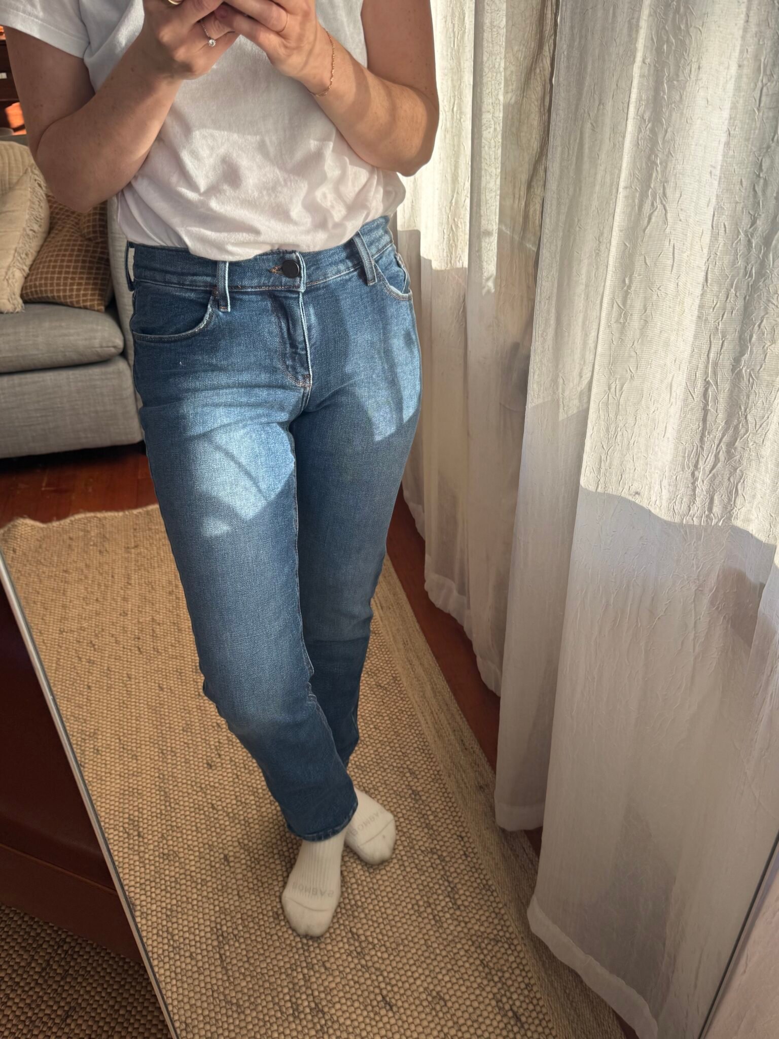 Person wearing a white t-shirt, blue jeans, and white socks stands in front of a mirror, taking a photo. Light-colored curtains and a woven rug are visible in the background.