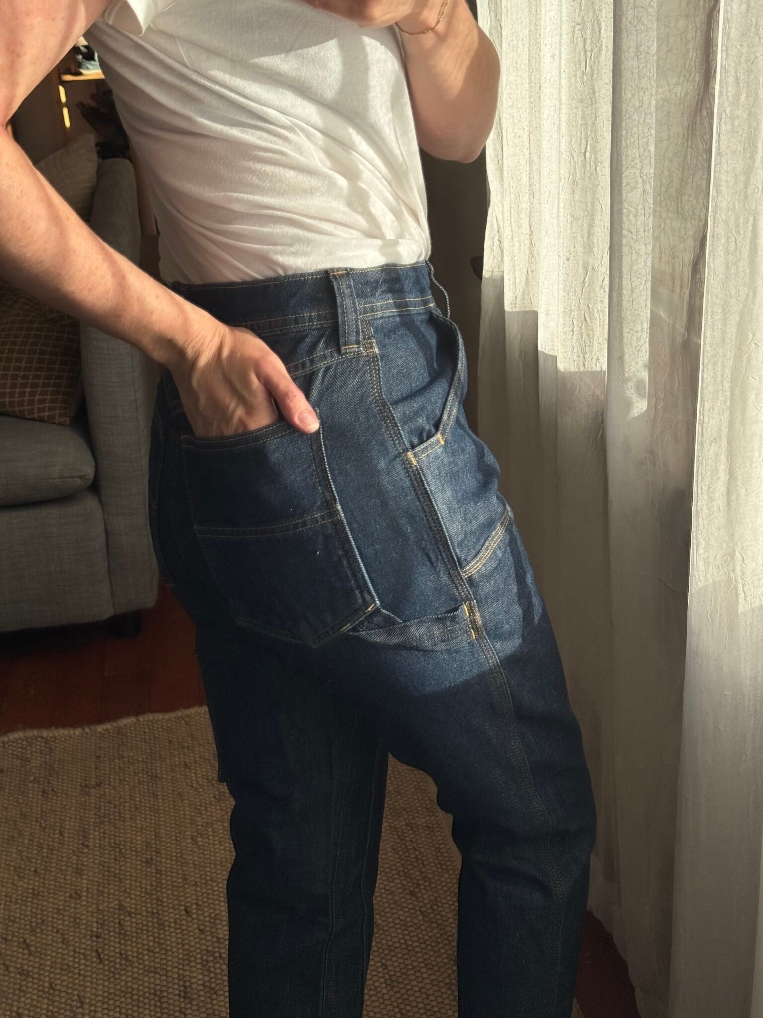 Person wearing a white t-shirt and blue jeans stands near a window, with one hand in the back pocket of the jeans. Natural light is coming through the curtains.