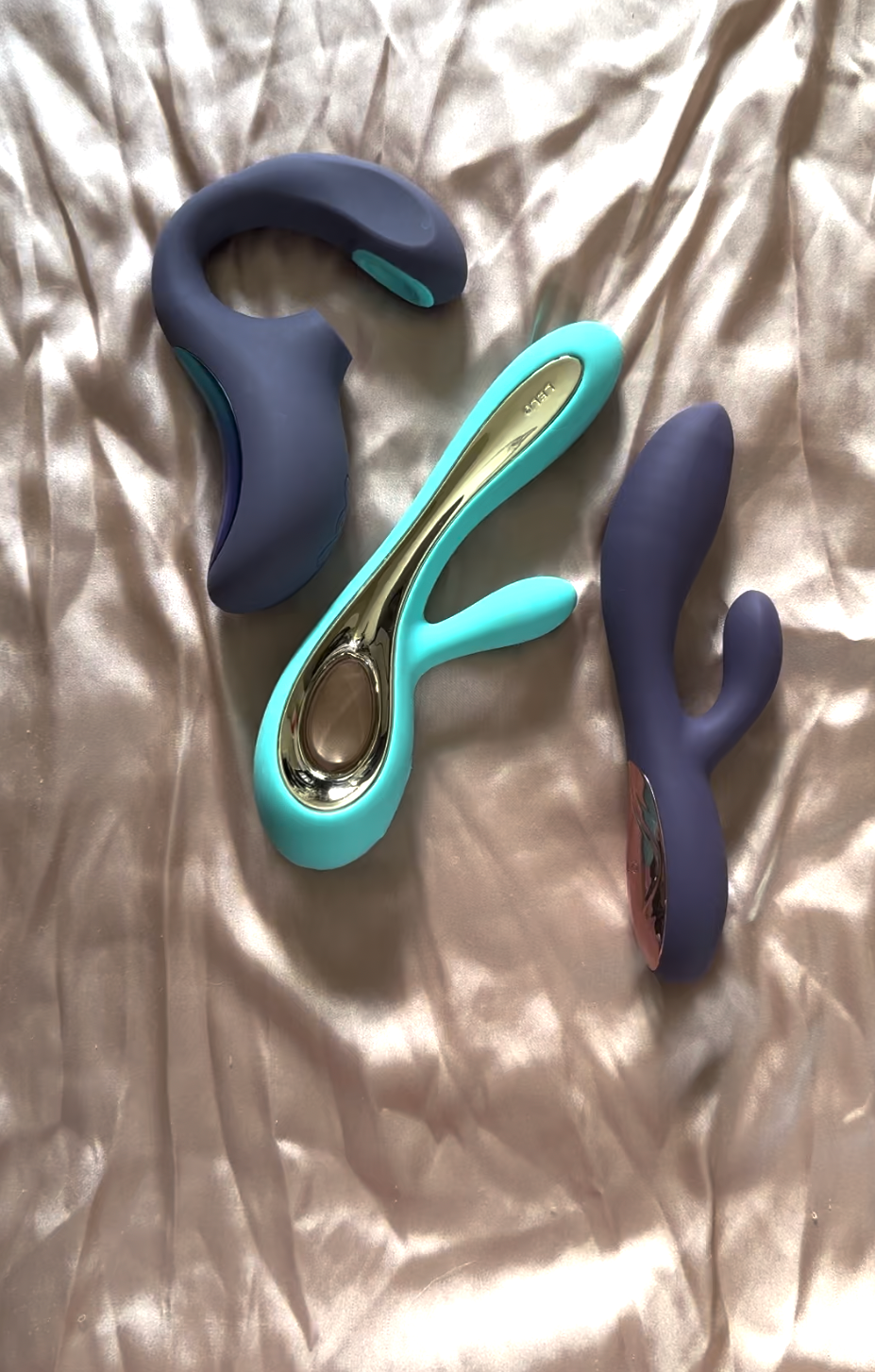 Three silicone vibrators in blue and teal colors rest on a silky, beige fabric background.