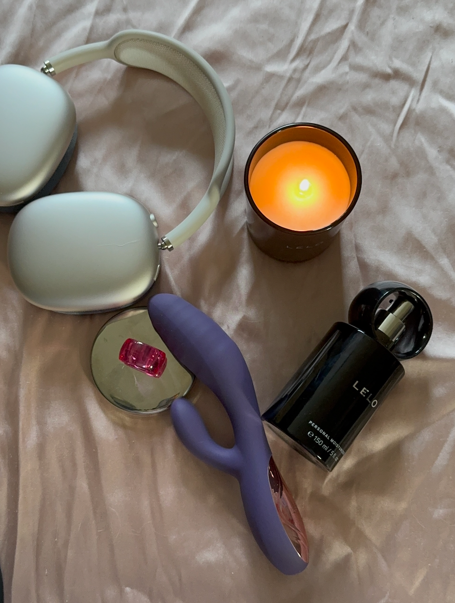 A pair of over-ear headphones, a lit candle, a bottle of LELO perfume, a purple vibrator, and a small pink accessory on a soft beige surface.