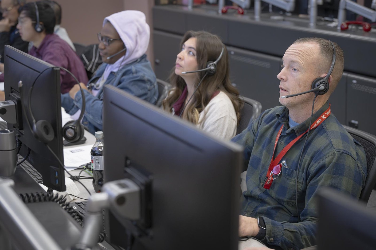 Six people sit at a long desk and face computer monitors. The person most in view, to the right of the frame, wears a green plaid button-down shirt and a red lanyard around his neck. Each person is wearing a headset with a microphone that connects to a computer.   