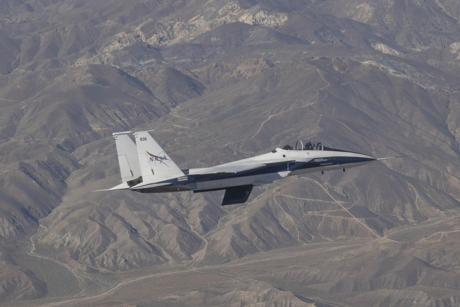 A white and blue painted jet aircraft flies above a mountain range. A model wing hangs below the aircraft’s center line. The cockpit is closed and two pilots are visible inside with flight gear on.