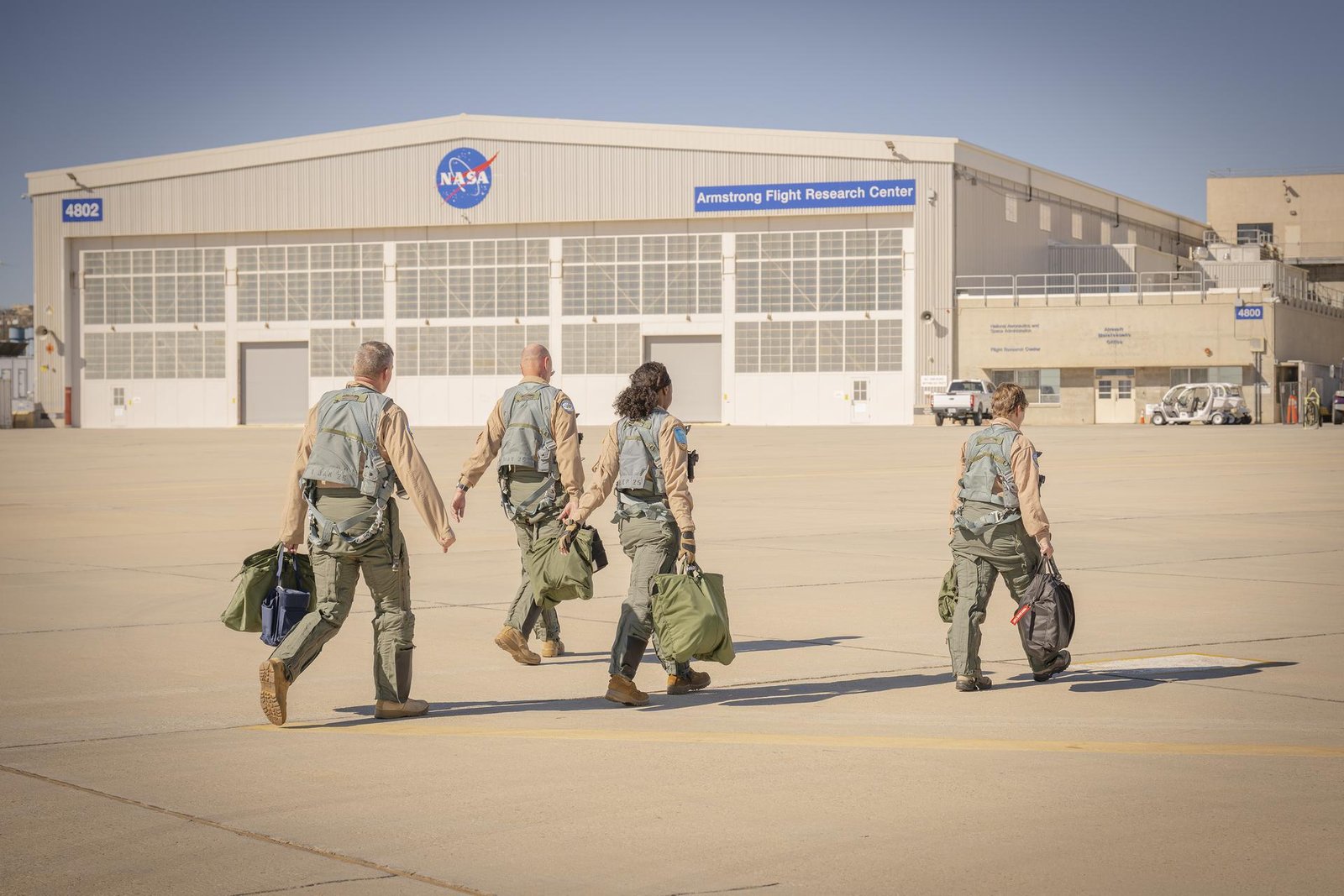 Four people walk toward a building on a concrete surface. Each person is wearing a flight harness, and other green flight gear, as well as a tan flight suit and tan boots. Each person also carries a flight helmet bag and other small bags with various flight gear inside.