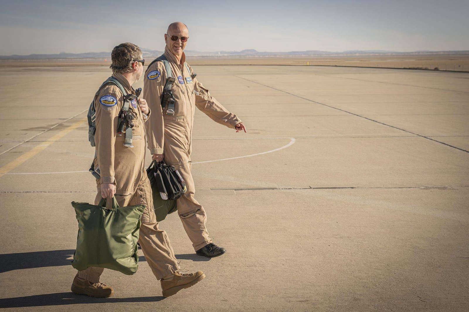 Two men wearing tan flight suits face each other and walk on a concrete surface. The men both carry pilot helmet bags with flight gear inside. Both men are wearing green flight gear.