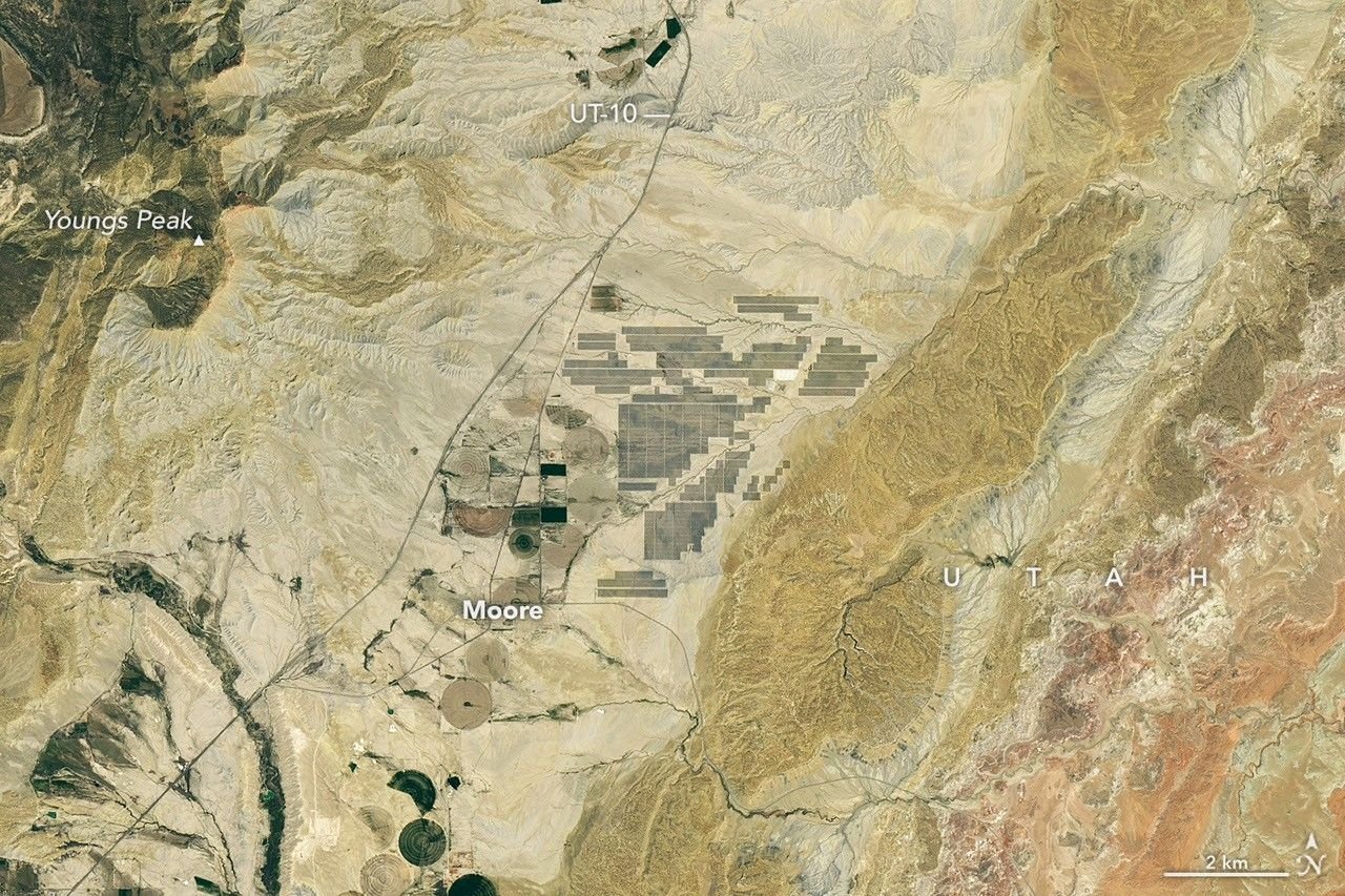 Grid-like arrays of solar panels cover parts of a valley floor in a brown, dry-looking area of Utah.