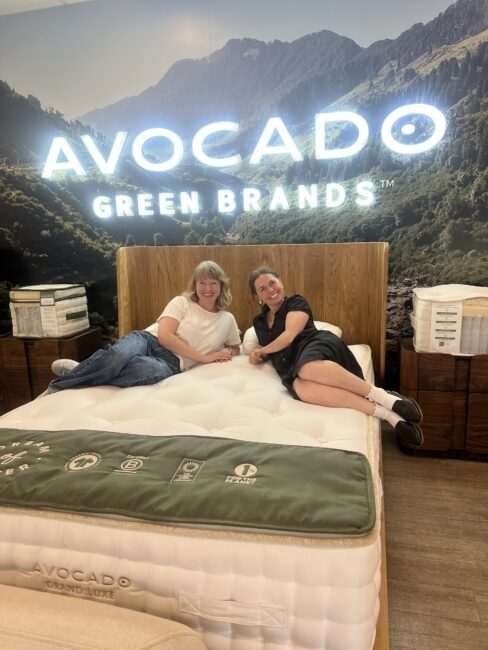 Two people sit and smile on an Avocado Green Brands mattress in a store, with a mountain mural and a branded sign in the background.