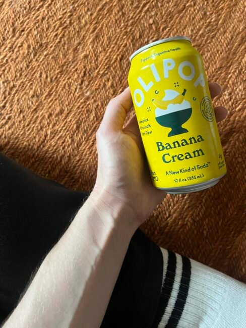 A hand holds a yellow can of OLIPOP Banana Cream soda over an orange textured surface, with part of a black pant leg and a white sock visible.