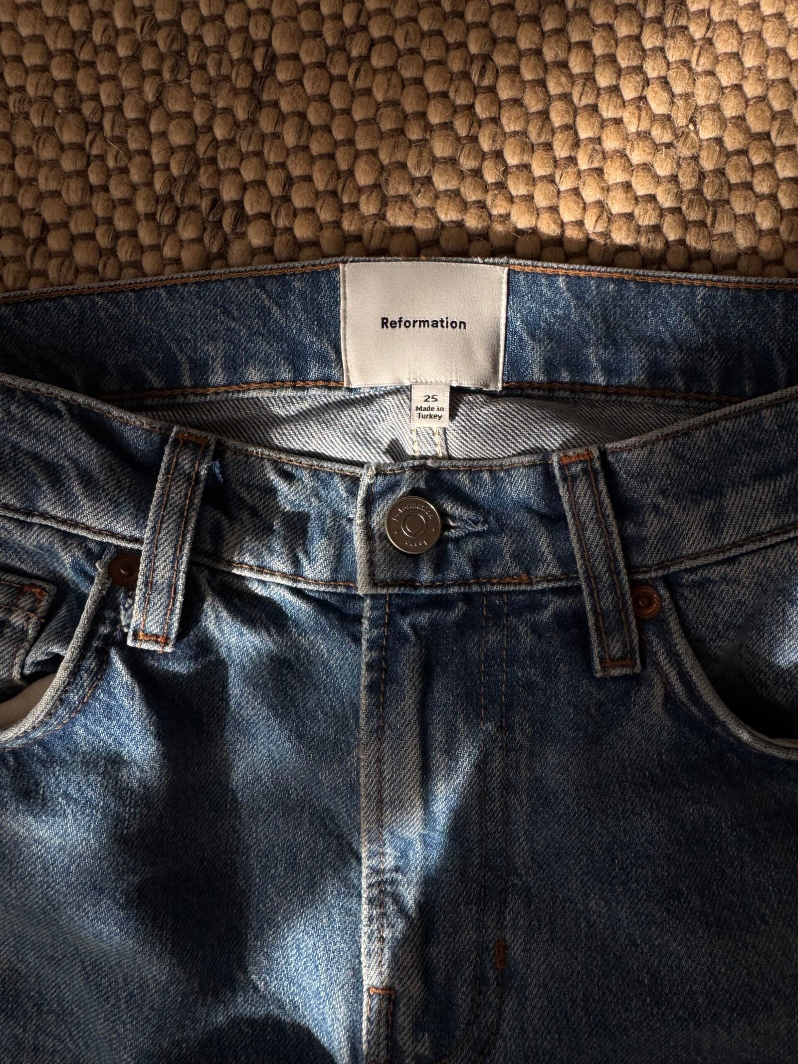 The waist area of blue Reformation jeans, size 25, made in Turkey, lying on a textured rug.